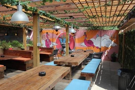 The Foundry's covered beer garden: long wooden tables and benches under a vine-covered pergola, with a painted flamingo mural along the back wall